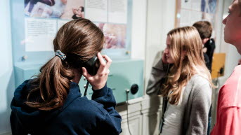 Två flickor i tonåren håller en hörlur intill örat och läser på textskyltar på en vägg.